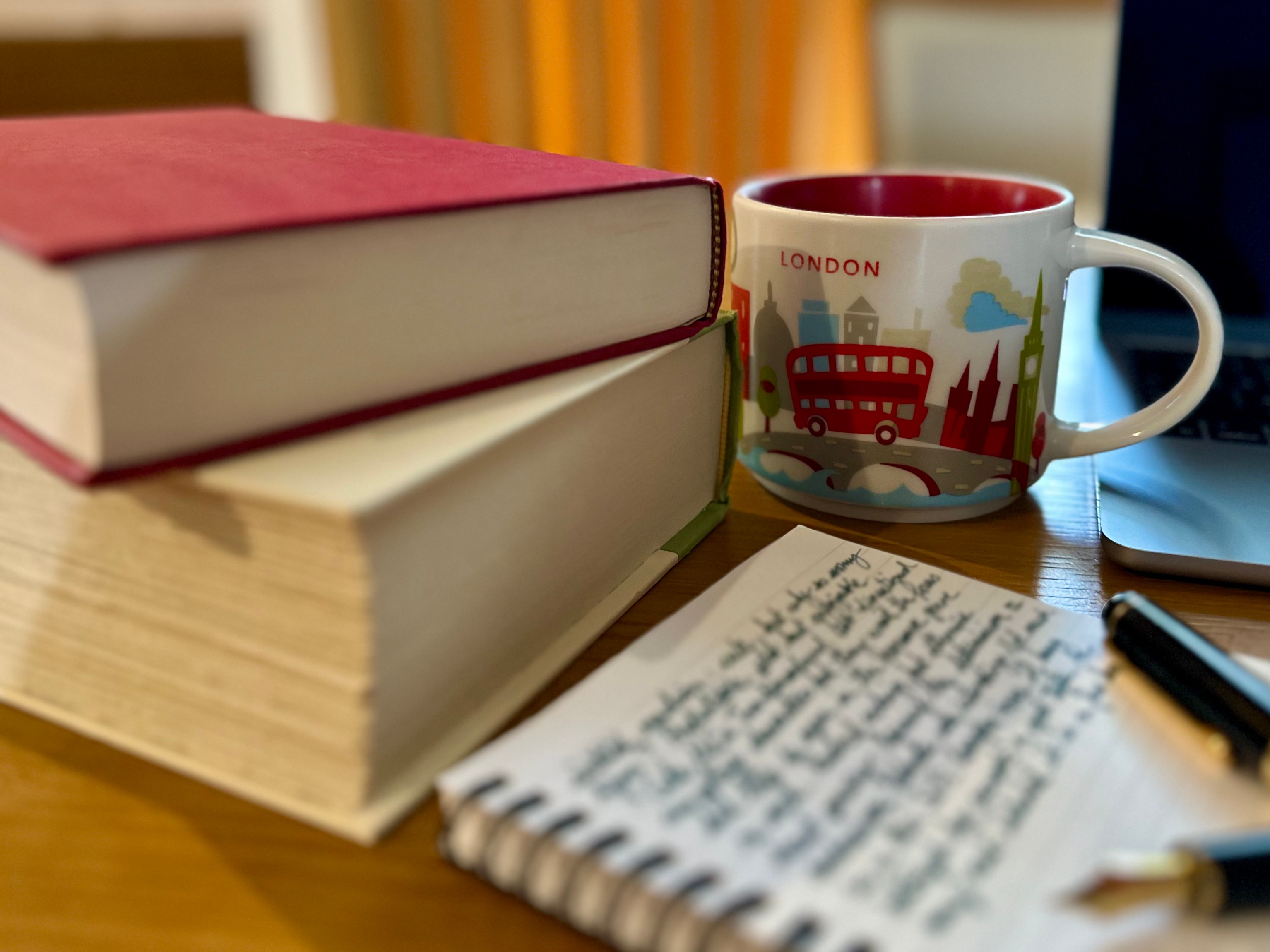 Two hardback books stacked on top of each other, next to a large mug of coffee decorated with images of London. In front of them both is a notebook on which black cursive writing is out of focus and thus illegible. 