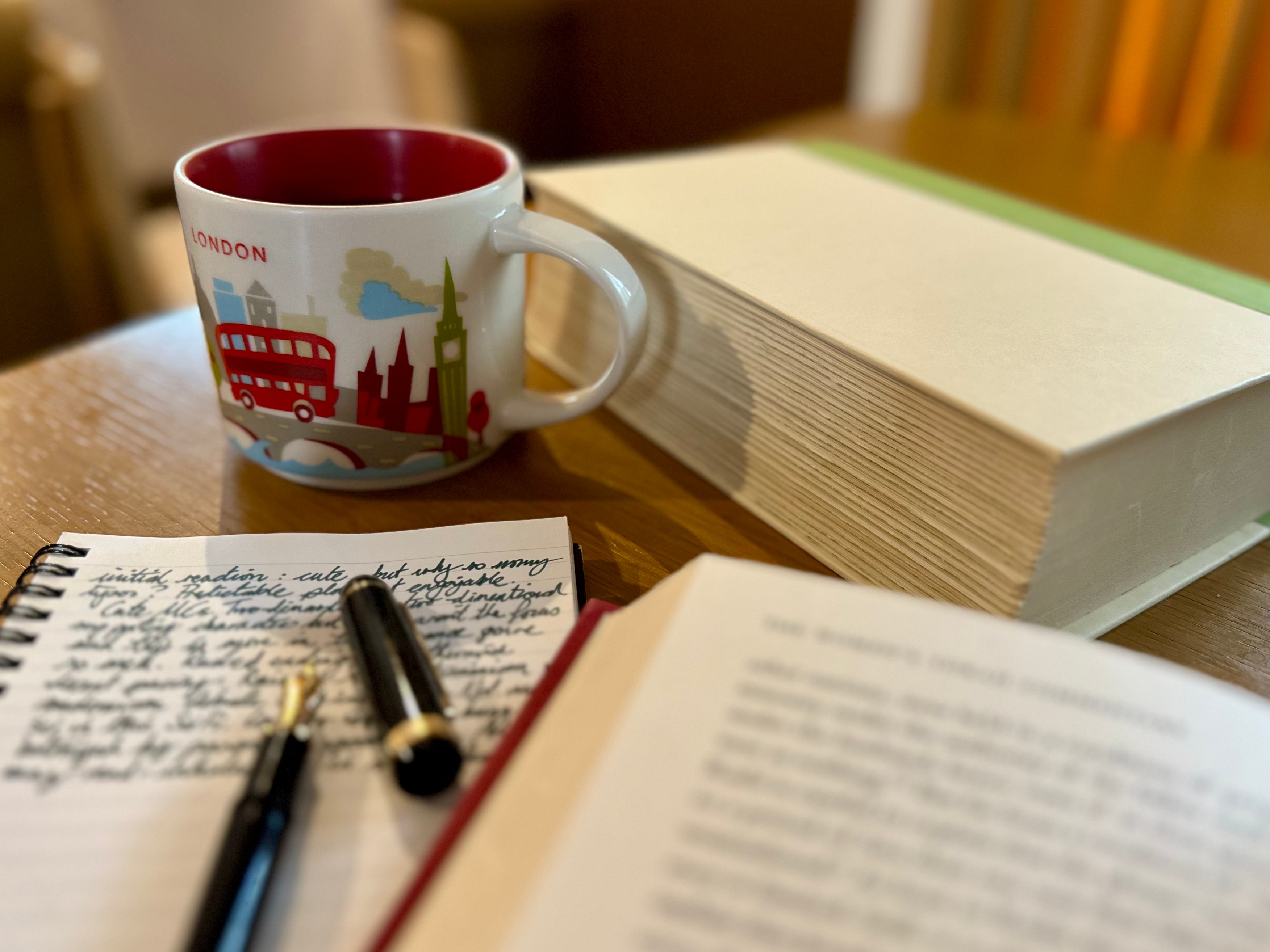 An open hardcover book, text out of focus, sits next to a spiral bound notebook and a fountain pen. All text is out of focus and thus illegible; the focus is instead on a large mug of coffee and a second hardback book in the background.